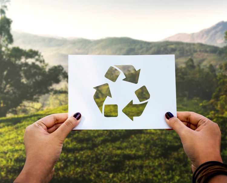 deux mains tenant un pochoir représentant le logo recyclage devant un paysage montagnard d'été